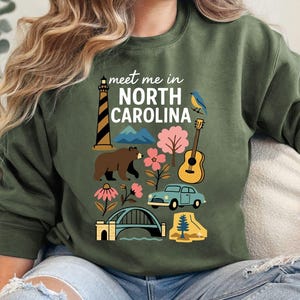 A woman wearing a green sweatshirt with a graphic design of various North Carolina landmarks and symbols, including a lighthouse, mountains, a bear, and a guitar.