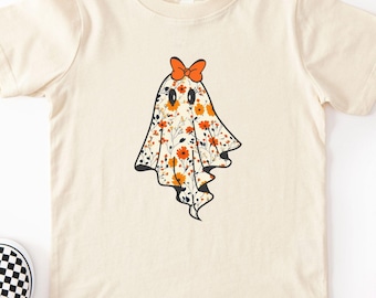 May include: Cream-colored t-shirt featuring a ghost design with a floral pattern in orange, white, and black. The ghost has an orange bow. Black and white checkered slip-on shoes are on the left. Sunglasses and crayons are on the right.