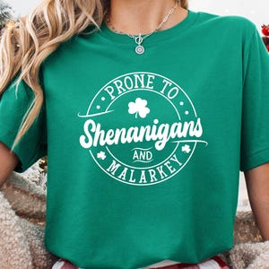A woman wearing a green t-shirt with white text and a shamrock graphic, standing in front of a Christmas tree.