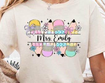 May include: Cream-colored t-shirt with a colorful pencil design and the text "Mrs. Emily". The pencils are decorated with bows and flowers. The word "NATURAL" is printed at the top left.