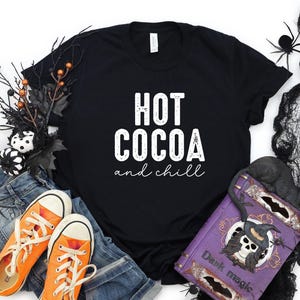a black t-shirt with the text "HOT COCOA and chill" printed on it, surrounded by Halloween-themed items such as a pair of orange sneakers, a pair of blue jeans, and a purple book with a skull and bat design.