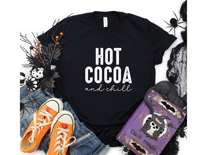 a black t-shirt with the text "HOT COCOA and chill" printed on it, surrounded by Halloween-themed items such as a pair of orange sneakers, a pair of blue jeans, and a purple book with a skull and bat design.