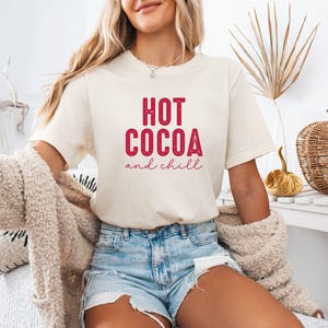 A woman wearing a white t-shirt with the text "HOT COCOA and chill" is sitting on a couch, with a beige sweater draped over her.