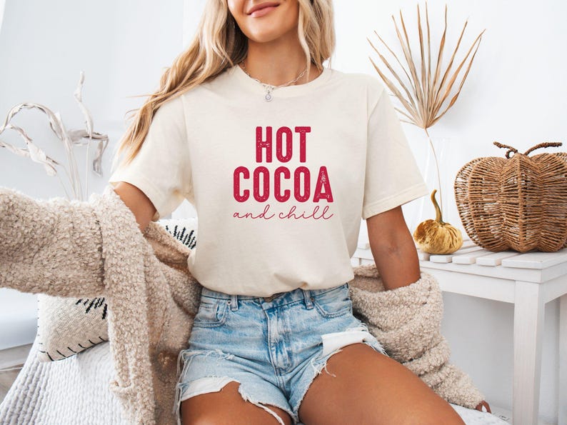 A woman wearing a white t-shirt with the text "HOT COCOA and chill" is sitting on a couch, with a beige sweater draped over her.