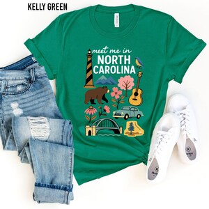 A green t-shirt with a graphic design featuring various elements associated with North Carolina, such as a lighthouse, a bear, a guitar, and a car, along with the text "Meet me in North Carolina". The t-shirt is laid out on a white background, accompanied by a pair of jeans and white sneakers.