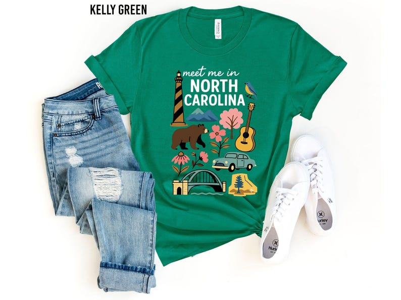 A green t-shirt with a graphic design featuring various elements associated with North Carolina, such as a lighthouse, a bear, a guitar, and a car, along with the text "Meet me in North Carolina". The t-shirt is laid out on a white background, accompanied by a pair of jeans and white sneakers.