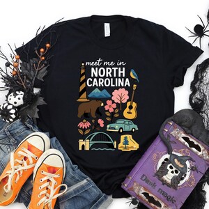 a black t-shirt with a graphic design of various North Carolina-themed items, including a lighthouse, a bear, a guitar, and a bridge. The t-shirt is surrounded by Halloween decorations, such as a spider web, a skull, and a bat. There are also some orange and purple sneakers, a purple book, and a black bag in the scene.