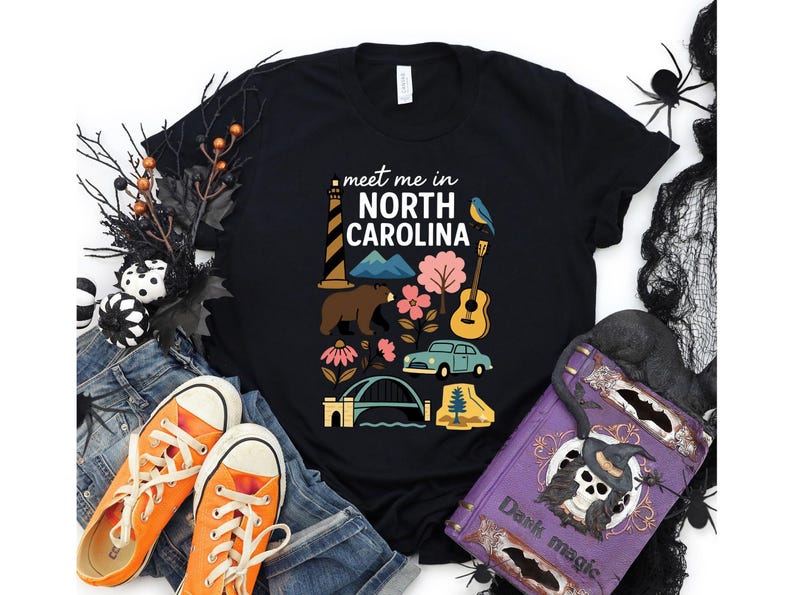 a black t-shirt with a graphic design of various North Carolina-themed items, including a lighthouse, a bear, a guitar, and a bridge. The t-shirt is surrounded by Halloween decorations, such as a spider web, a skull, and a bat. There are also some orange and purple sneakers, a purple book, and a black bag in the scene.