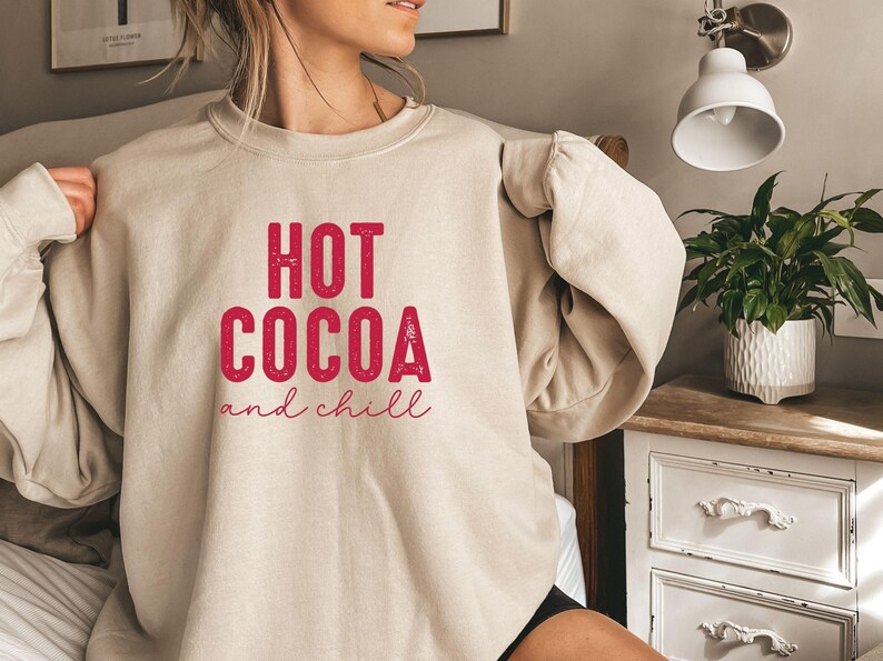A woman wearing a beige sweatshirt with the text "HOT COCOA and chill" printed on it, standing in a bedroom with a potted plant and a dresser visible in the background.