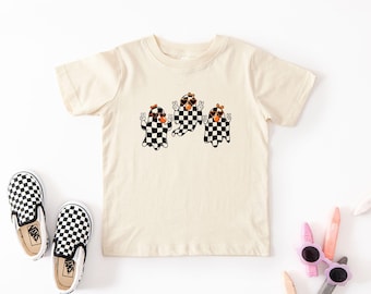 May include: Cream-colored t-shirt featuring three cartoon ghosts with black and white checkered patterns. The ghosts are wearing sunglasses and bows, with peace signs. Checkered slip-on shoes are on the left. A fun, playful design for kids.