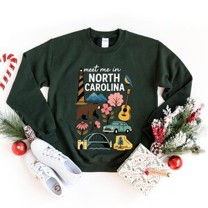 A green sweatshirt with a graphic design featuring various North Carolina-related icons, such as a lighthouse, pine tree, and car, surrounded by Christmas decorations and gifts.