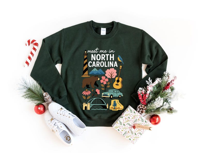 A green sweatshirt with a graphic design featuring various North Carolina-related icons, such as a lighthouse, pine tree, and car, surrounded by Christmas decorations and gifts.