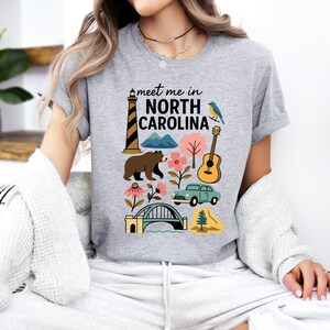 A woman wearing a gray t-shirt with a graphic design featuring various elements associated with North Carolina, such as a lighthouse, bear, guitar, and bridge.