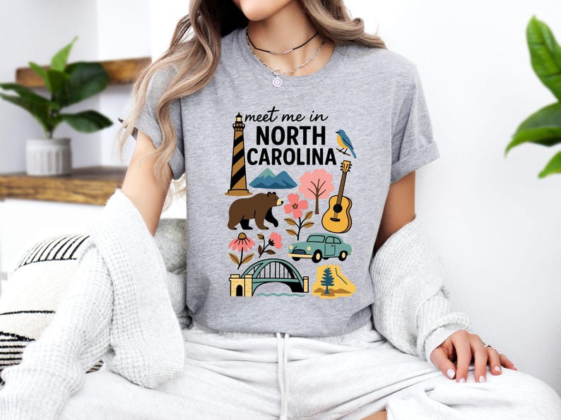 A woman wearing a gray t-shirt with a graphic design featuring various elements associated with North Carolina, such as a lighthouse, bear, guitar, and bridge.