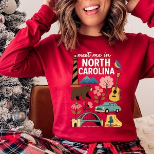A woman wearing a red long-sleeved shirt with a graphic design featuring various North Carolina landmarks and symbols, including a lighthouse, mountains, a bear, a guitar, and a car.