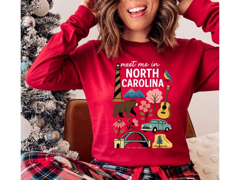 A woman wearing a red long-sleeved shirt with a graphic design featuring various North Carolina landmarks and symbols, including a lighthouse, mountains, a bear, a guitar, and a car.