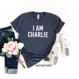 a casual outfit consisting of a gray t-shirt with the text "I AM CHARLIE", a pair of blue jeans, white sneakers, and a bouquet of pink flowers.