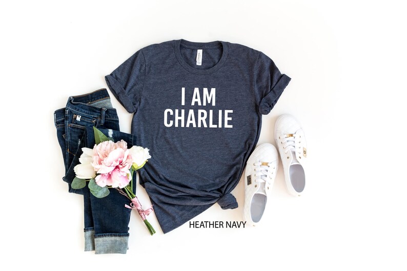 a casual outfit consisting of a gray t-shirt with the text "I AM CHARLIE", a pair of blue jeans, white sneakers, and a bouquet of pink flowers.