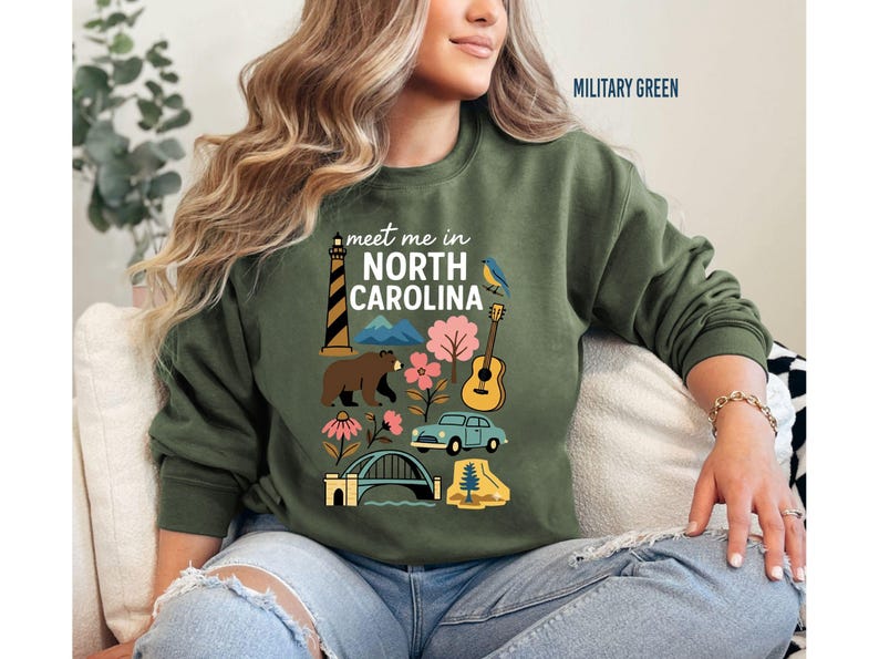 A woman wearing a green sweatshirt with a graphic design of various North Carolina landmarks and symbols, including a lighthouse, mountains, a bear, and a guitar.