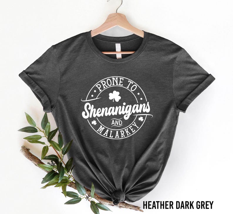 A black t-shirt with a white graphic design that says "PRONTO SHENANIGANS AND MALARKEY" printed on it, hanging on a wooden hanger.