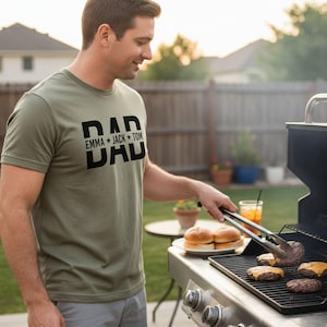 A man in a green t-shirt is grilling hamburgers and hot dogs on a barbecue grill in a backyard setting.