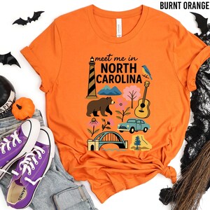 a t-shirt with a graphic design of various North Carolina landmarks and symbols, including a lighthouse, a bear, a guitar, and a car, along with the text "Meet me in North Carolina". The t-shirt is displayed on a white background, surrounded by Halloween-themed items such as bats, a broom, and a pumpkin.