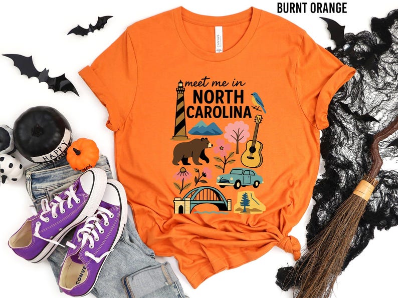 a t-shirt with a graphic design of various North Carolina landmarks and symbols, including a lighthouse, a bear, a guitar, and a car, along with the text "Meet me in North Carolina". The t-shirt is displayed on a white background, surrounded by Halloween-themed items such as bats, a broom, and a pumpkin.