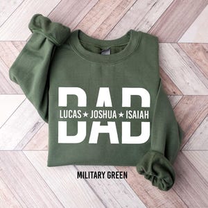 A green sweatshirt with the text "DAD" printed on it, along with the names "LUCAS", "JOSHUA", and "ISAIAH". The sweatshirt is laid out on a wooden surface.