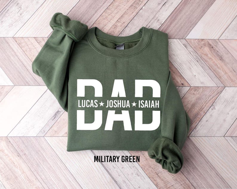 A green sweatshirt with the text "DAD" printed on it, along with the names "LUCAS", "JOSHUA", and "ISAIAH". The sweatshirt is laid out on a wooden surface.