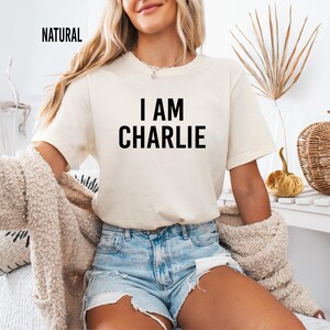 A woman wearing a white t-shirt with the text "I AM CHARLIE" and denim shorts is sitting on a couch, with a woven basket and a plant in the background.