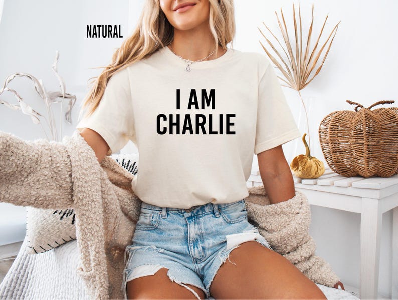 A woman wearing a white t-shirt with the text "I AM CHARLIE" and denim shorts is sitting on a couch, with a woven basket and a plant in the background.
