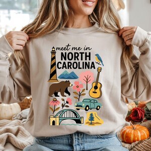A woman wearing a beige sweater with a graphic design of various North Carolina landmarks and symbols, including a lighthouse, mountains, a bear, a guitar, and a pumpkin.