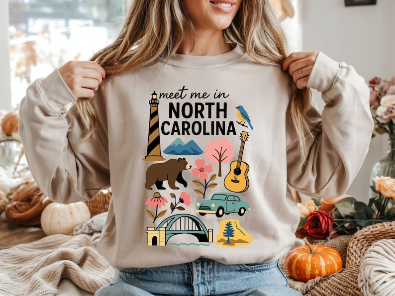 A woman wearing a beige sweater with a graphic design of various North Carolina landmarks and symbols, including a lighthouse, mountains, a bear, a guitar, and a pumpkin.