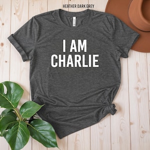 a gray t-shirt with the text "I AM CHARLIE" printed on it, along with a brown hat and a potted plant on a wooden surface.