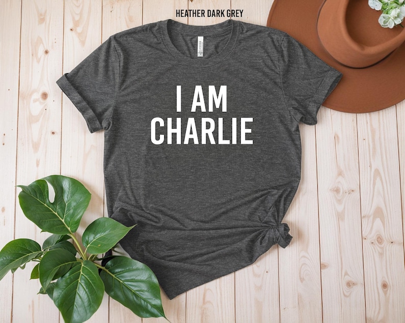 a gray t-shirt with the text "I AM CHARLIE" printed on it, along with a brown hat and a potted plant on a wooden surface.