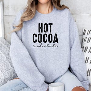 A woman wearing a grey sweatshirt with the text "HOT COCOA and chill" printed on it, sitting on a couch with a patterned pillow behind her.