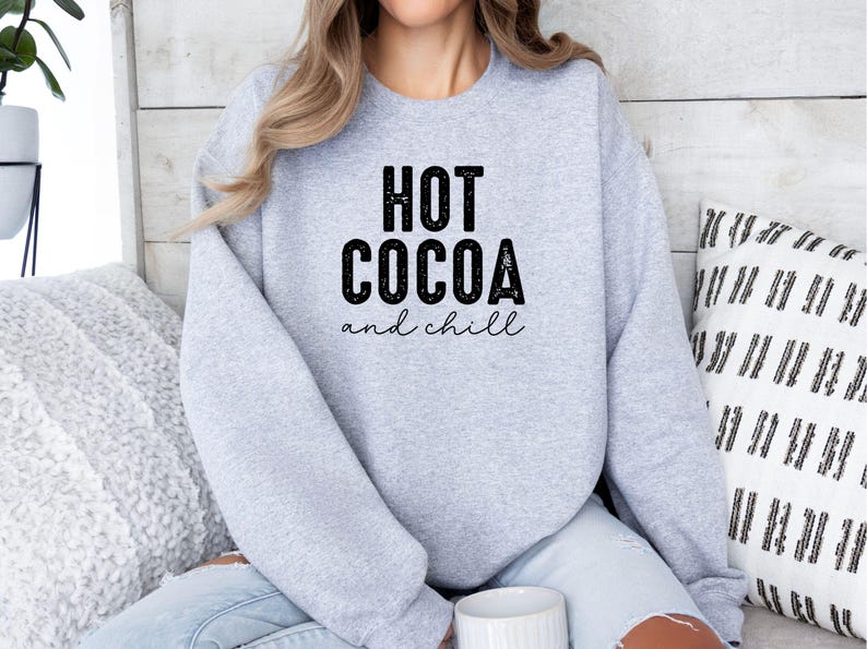 A woman wearing a grey sweatshirt with the text "HOT COCOA and chill" printed on it, sitting on a couch with a patterned pillow behind her.