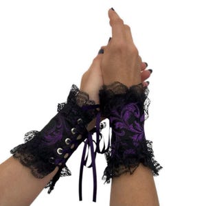 Puede incluir: Puños bordados en negro y morado con ribete de encaje. Los puños presentan un diseño floral y están atados con una cinta morada. Los puños se llevan en las muñecas.