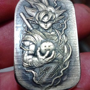 May include: A rectangular, silver-toned pendant with a detailed relief of a Dragon Ball Z character. The character is holding a glowing orb. The pendant has a textured background and intricate design details.