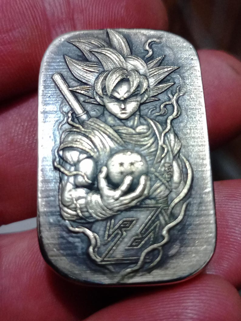 May include: A rectangular, silver-toned pendant with a detailed relief of a Dragon Ball Z character. The character is holding a glowing orb. The pendant has a textured background and intricate design details.