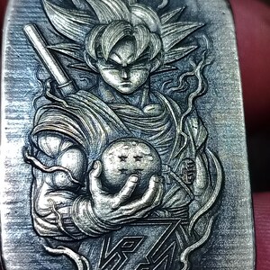 May include: A silver pendant with a detailed relief of a character with spiky hair, holding a sphere with stars. The pendant also includes the stylized text "Z". The design is intricate and detailed, with a focus on the character's muscular physique.