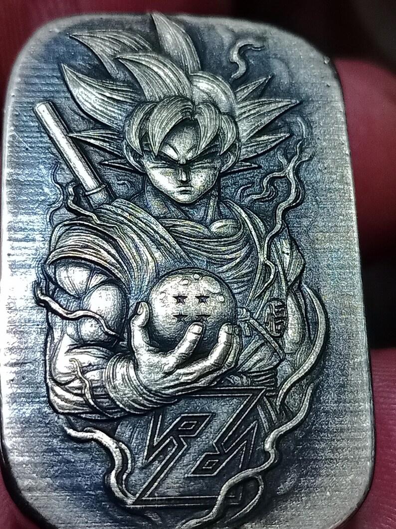 May include: A silver pendant with a detailed relief of a character with spiky hair, holding a sphere with stars. The pendant also includes the stylized text "Z". The design is intricate and detailed, with a focus on the character's muscular physique.