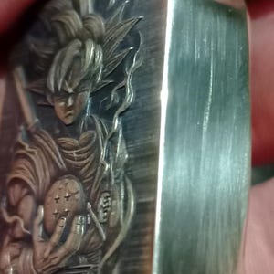 May include: A silver metal lighter with an engraved image of a character holding a dragon ball. The design includes detailed depictions of the character's hair, clothing, and the dragon ball. The lighter has a brushed metal finish and a rectangular shape.