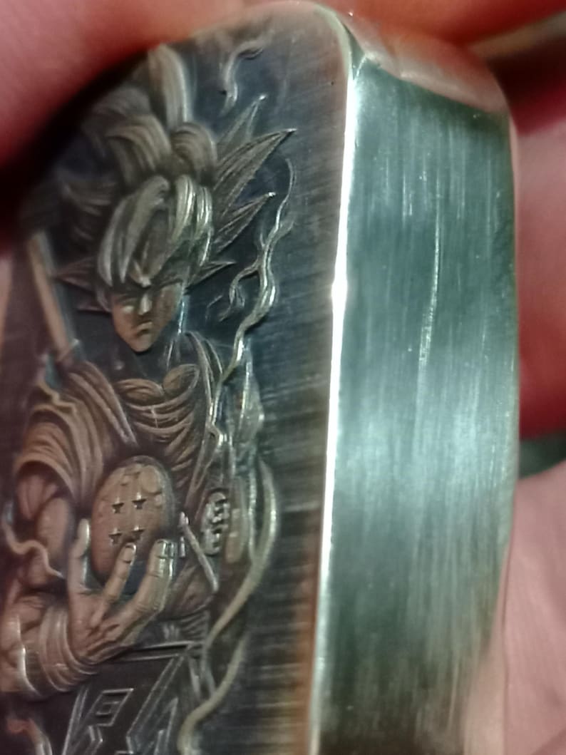 May include: A silver metal lighter with an engraved image of a character holding a dragon ball. The design includes detailed depictions of the character's hair, clothing, and the dragon ball. The lighter has a brushed metal finish and a rectangular shape.