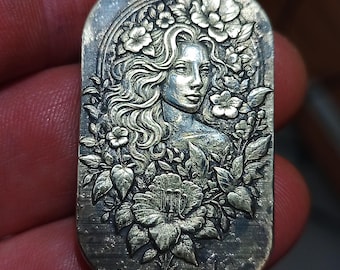 May include: A rectangular, silver-coloured metal pendant with a detailed relief of a woman's face framed by flowers and foliage. The woman has long, flowing hair and a calm expression. The floral design includes various types of blossoms and leaves.