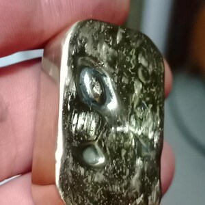 May include: A silver ring with a rectangular face. The face has a textured, organic appearance with a dark green hue. The ring is held between fingers, showcasing its polished metal and unique design.