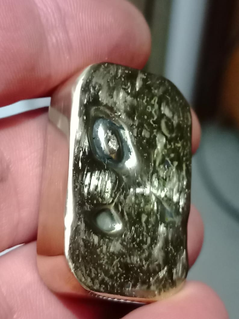 May include: A silver ring with a rectangular face. The face has a textured, organic appearance with a dark green hue. The ring is held between fingers, showcasing its polished metal and unique design.
