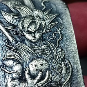 May include: A silver-coloured pendant with a detailed relief of a Dragon Ball Z character. The character is depicted holding a dragon ball with stars. The pendant has intricate detailing and a textured finish.