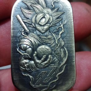 May include: A rectangular, silver-toned pendant with a detailed relief of a character with spiky hair, holding a glowing orb. The character is from the Dragon Ball Z series. The pendant has a textured surface and intricate design.