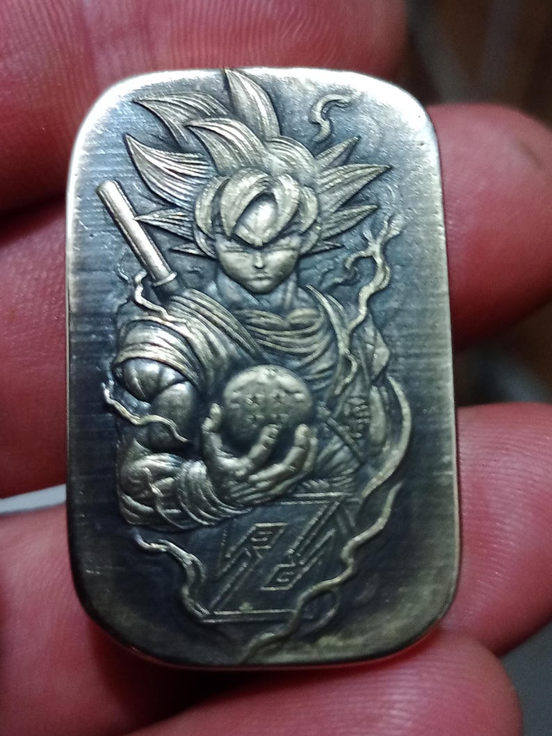 May include: A rectangular, silver-toned pendant with a detailed relief of a character with spiky hair, holding a glowing orb. The character is from the Dragon Ball Z series. The pendant has a textured surface and intricate design.
